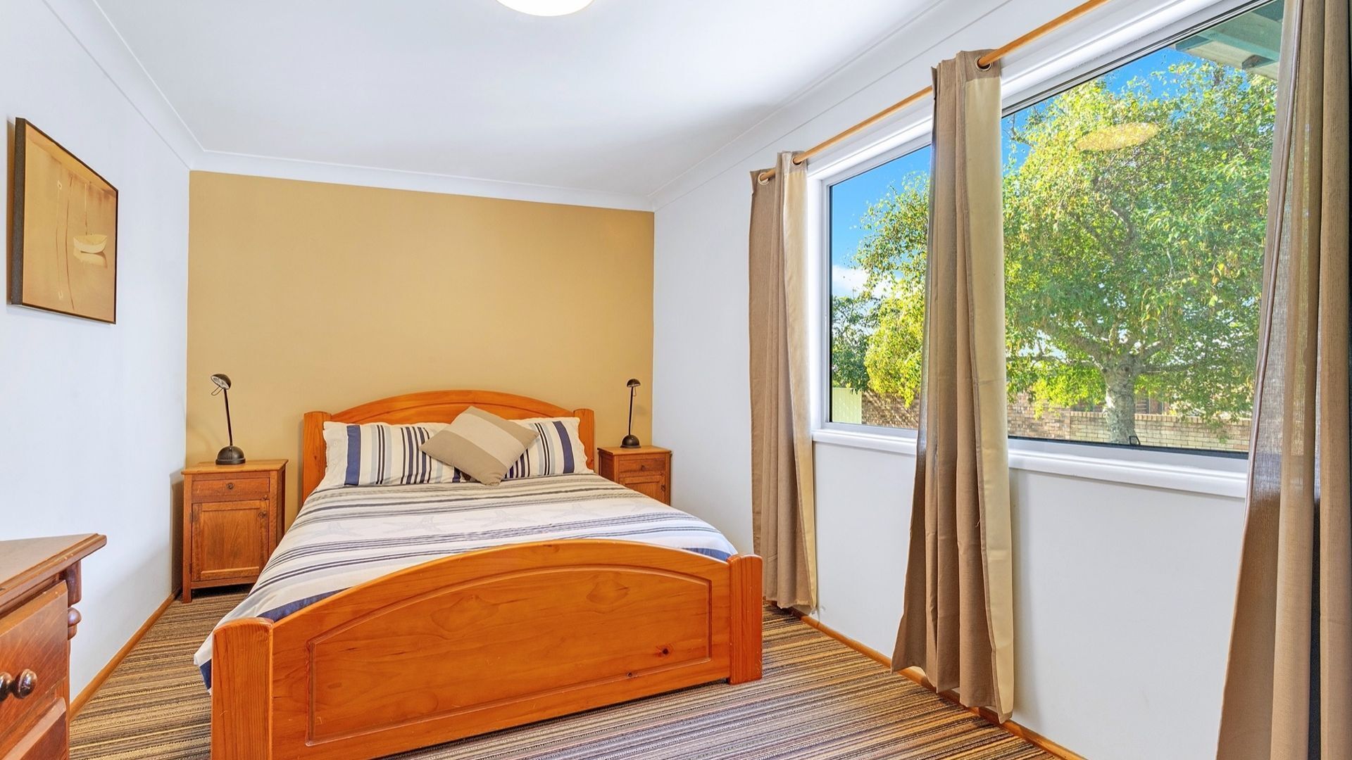 A bedroom with a wooden bed, two bedside tables, a nightstand, a window with curtains, and a painting on the wall.