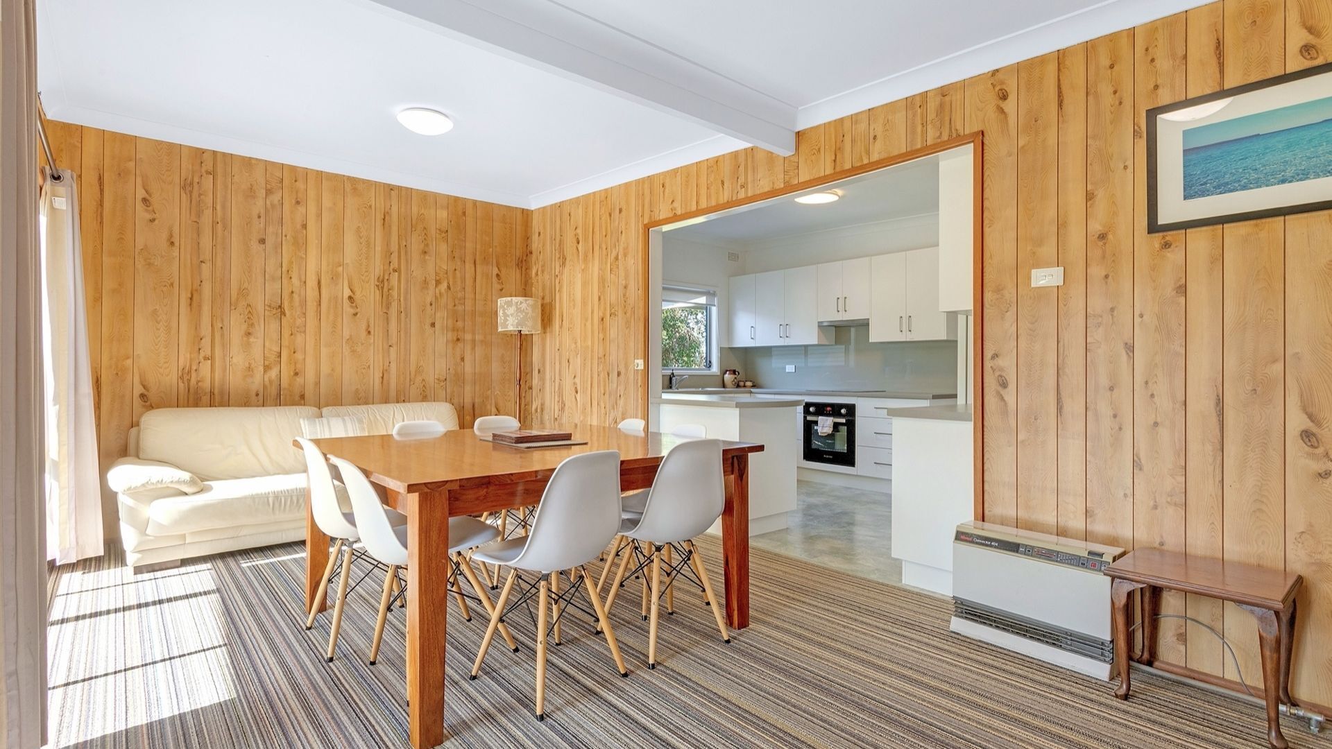 A cozy room with a wooden wall, a white sofa, a dining table with chairs, and a kitchen area visible through an open doorway.