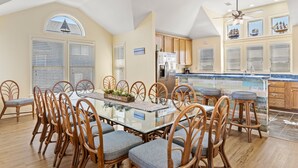 Un comedor espacioso con una mesa de vidrio rodeada de sillas de mimbre, una cocina con armarios de madera y un amplio ventanal con persianas.