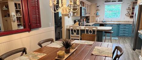 Un coin repas avec une table en bois, des chaises et un lustre. On aperçoit une cuisine en arrière-plan, avec des meubles bleus et une fenêtre.