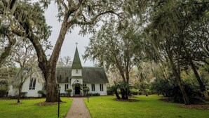 Une église blanche surmontée d’un clocher vert, entourée de grands arbres et bordée d’un chemin en briques.