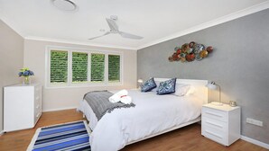 Une chambre à coucher comprenant un lit, des tables de chevet, un bureau, une fenêtre avec des stores, un ventilateur de plafond et un élément décoratif mural.