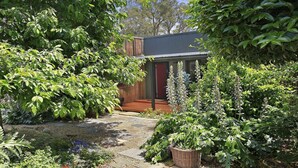 En havegang, der fører til en træterrasse med et metal tag, omgivet af frodig vegetation og forskellige planter.