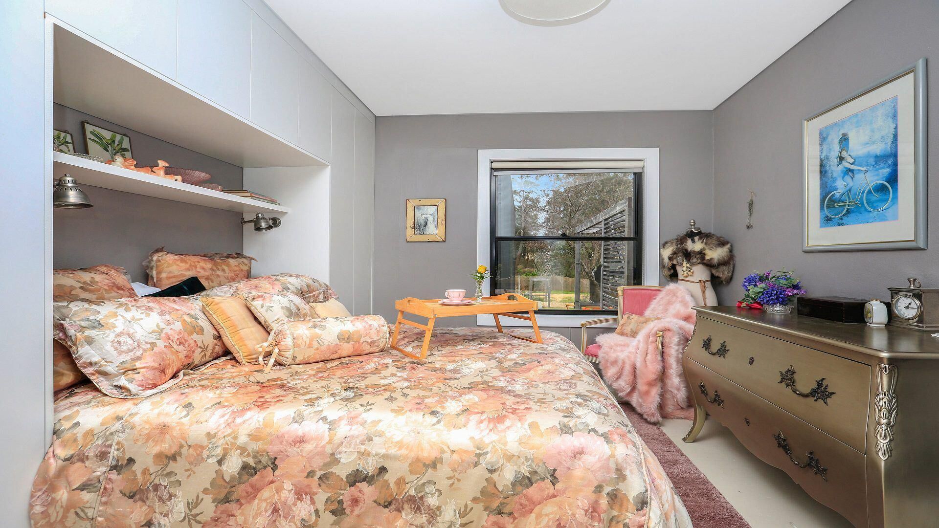 Quarto com colcha florida, cômoda, cadeira e janela com vista para árvores.