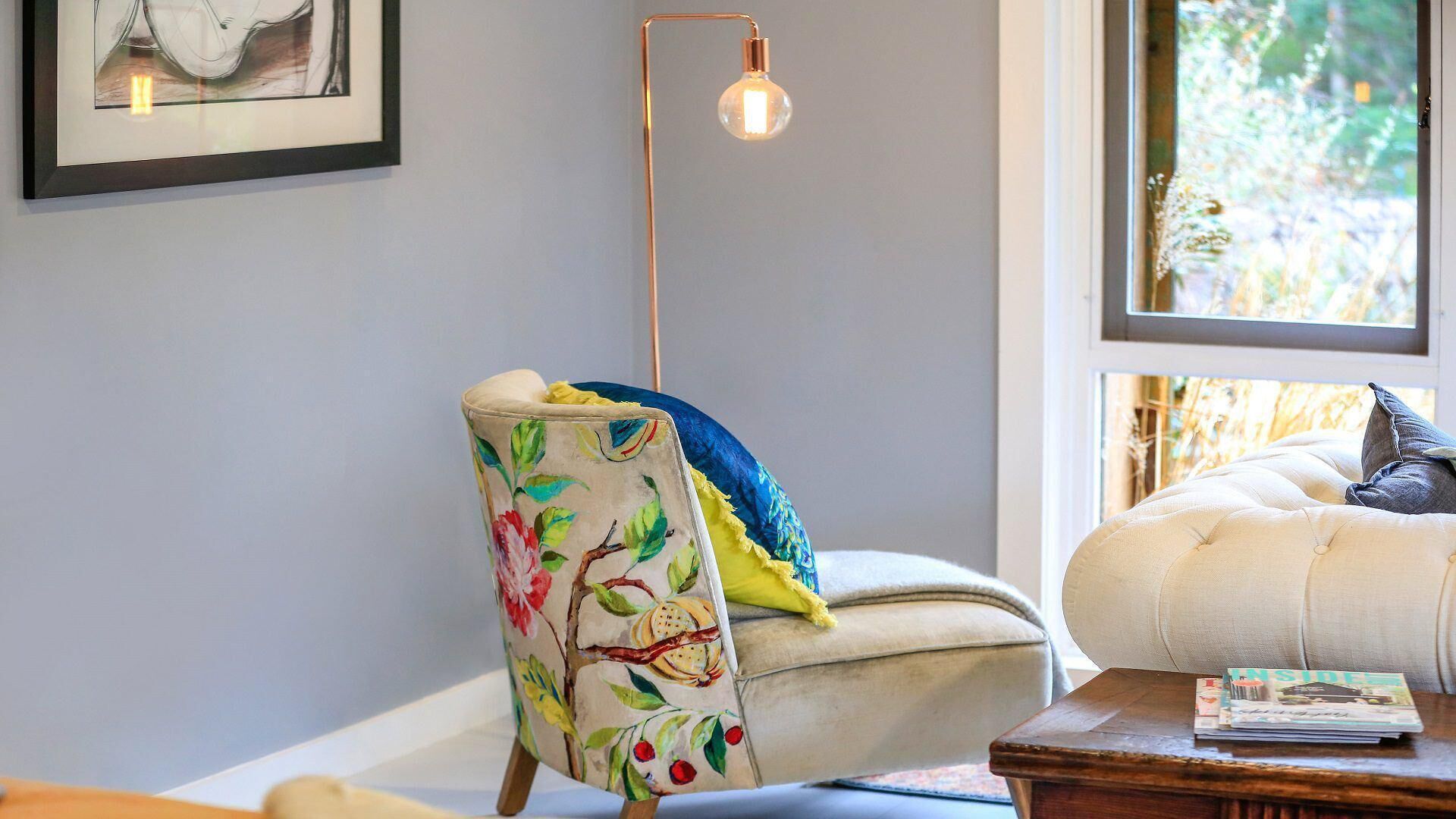Un salon chaleureux avec un fauteuil à motifs floraux, un lampadaire moderne et une fenêtre donnant sur un paysage verdoyant.