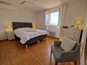 Une chambre à coucher comprenant un lit, des tables de chevet, une chaise, une commode, une fenêtre avec des rideaux et un radiateur.