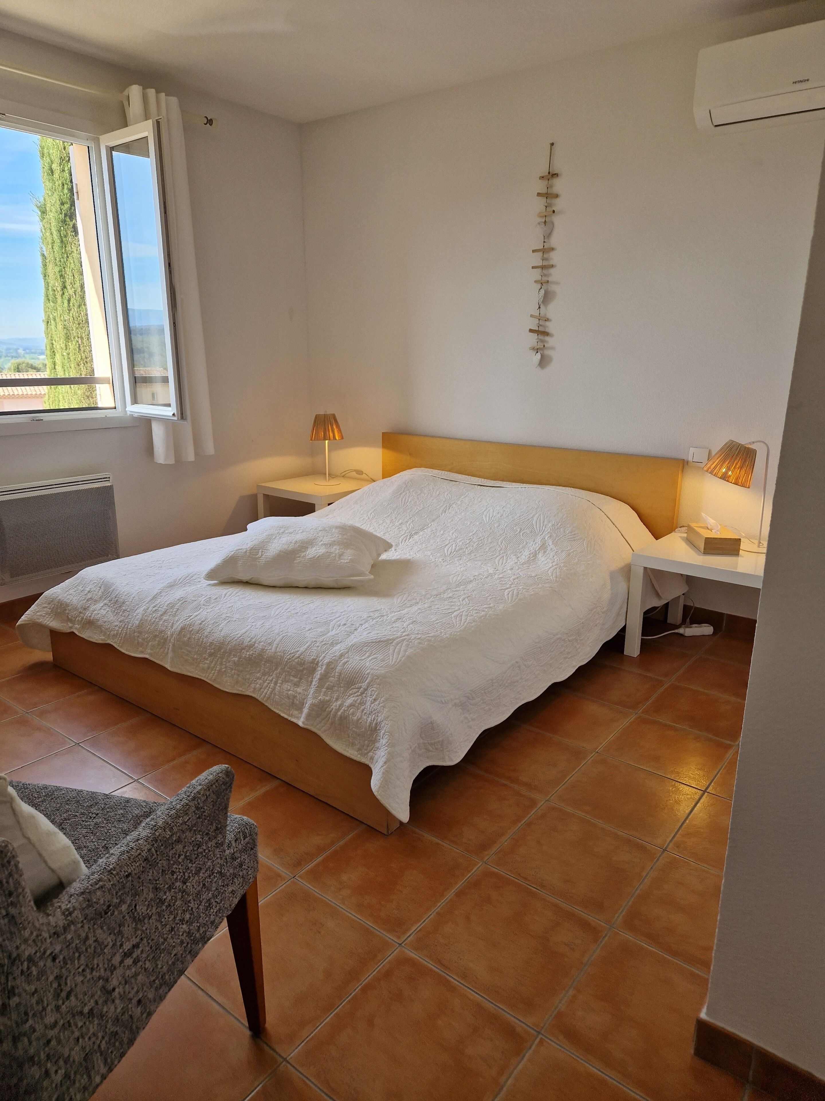 A bedroom with a bed, two bedside tables, a chair, a window with curtains, and an air conditioning unit.