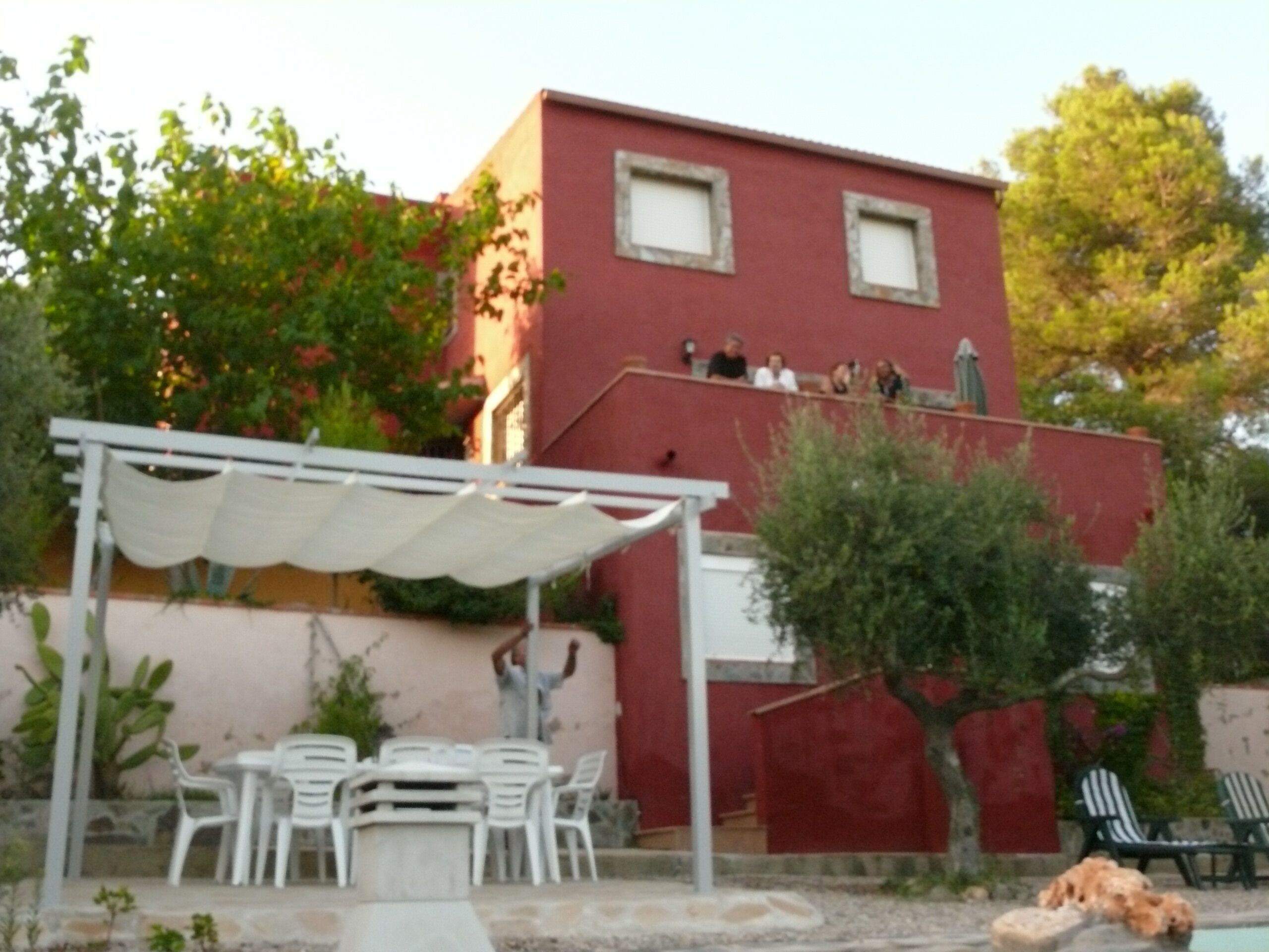 Un bâtiment rouge avec deux fenêtres, un patio couvert avec des chaises blanches et un petit espace piscine où se trouve un chien.