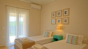 A hotel room with two beds, a window with sheer curtains, a wall-mounted air conditioner, and framed pictures above the beds. - Beach Villa Joanna - Luxury Villa rental in Corfu, Greece (Acharavi)