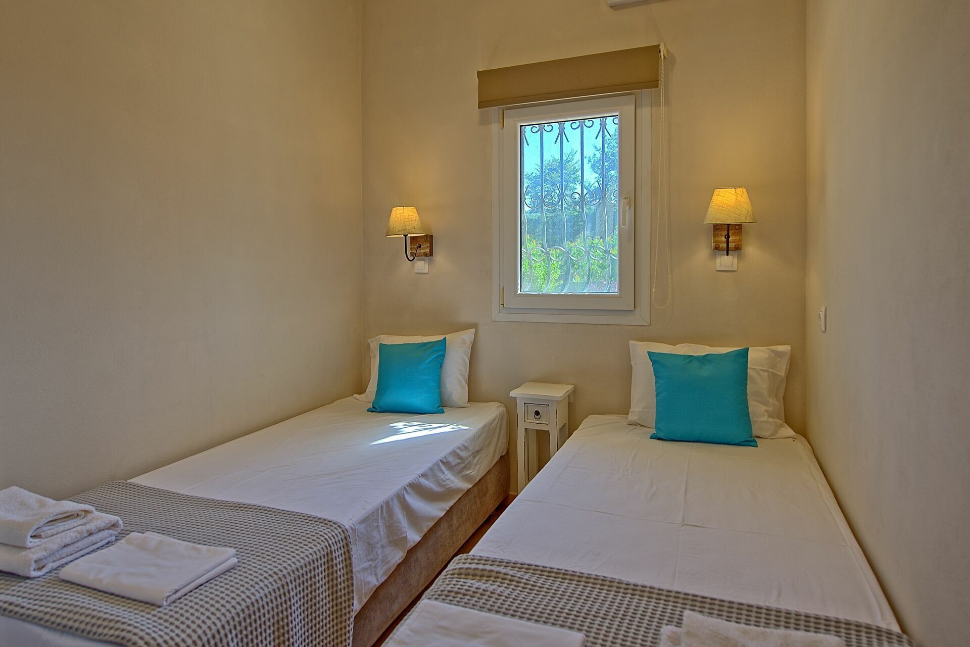 Ein Zimmer mit zwei Einzelbetten, beide mit weißer Bettdecke und blauem Kissen. An jeder Seite eines Fensters befindet sich eine Wandlampe.
