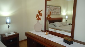 Una camera d'albergo con un comò in legno, uno specchio, un vaso con fiori, un comodino con una lampada e un letto con testiera.