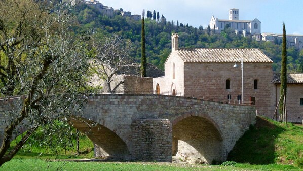 A stone bridge with three arches over a small stream, surrounded by greenery and a hill with buildings in the background.