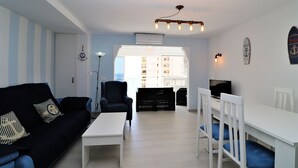 A living room with a black sofa, a white coffee table, a blue armchair, and a dining area with a white table and blue chairs. - Baccana 9-4 (Benidorm)