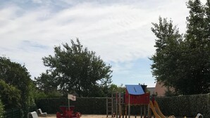 Un espace extérieur sablonneux avec une aire de jeux, comprenant un toboggan et des structures d’escalade, entouré de bancs et d’une haie.
