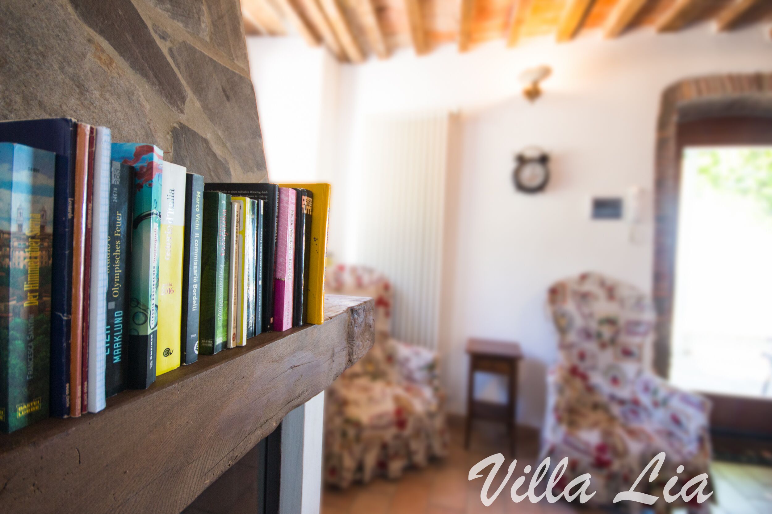 A bookshelf with books in a room with a floral armchair and a stone wall.