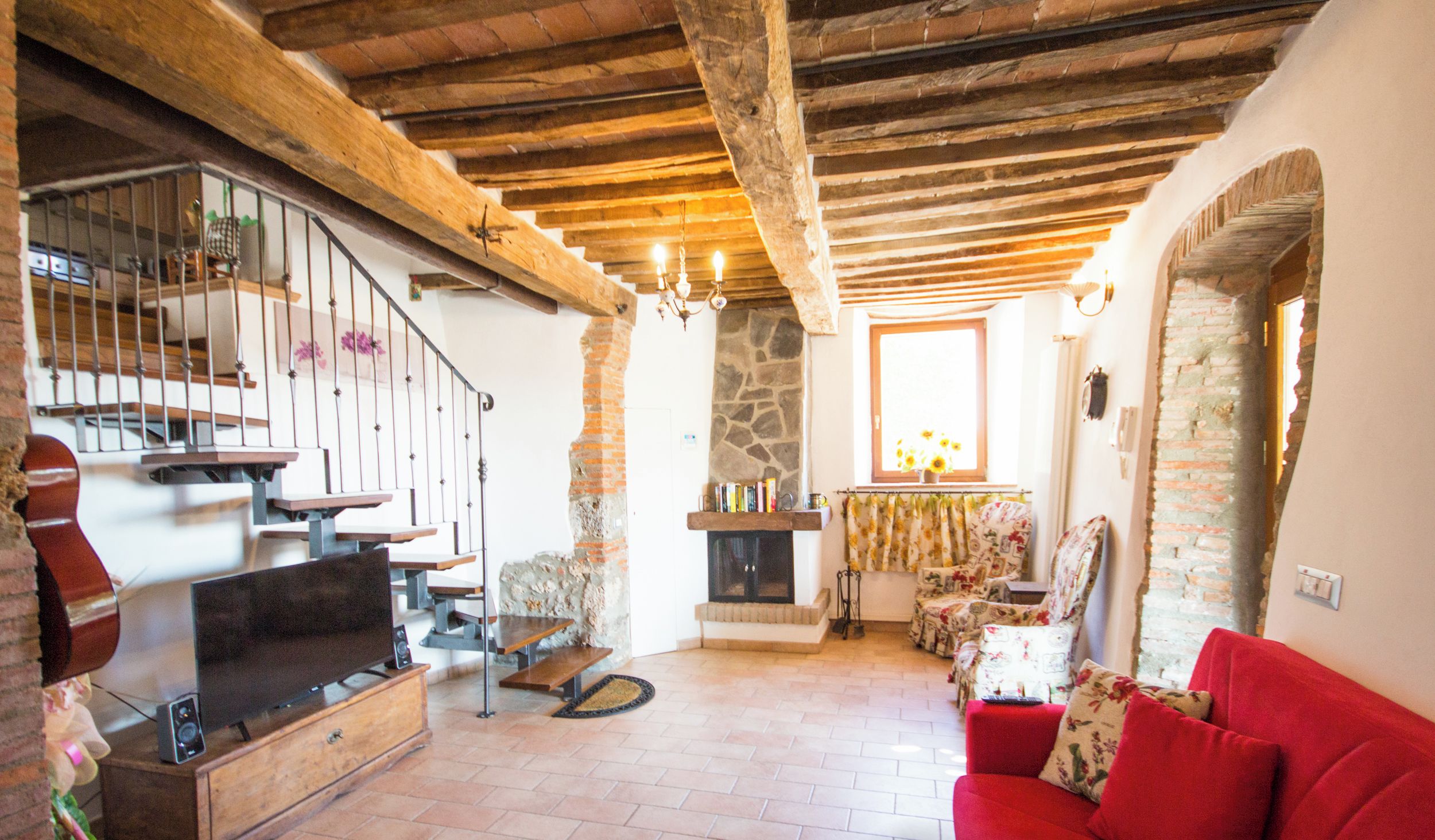 A cozy living room with a fireplace, wooden beams, a red sofa, and a television on a wooden cabinet.