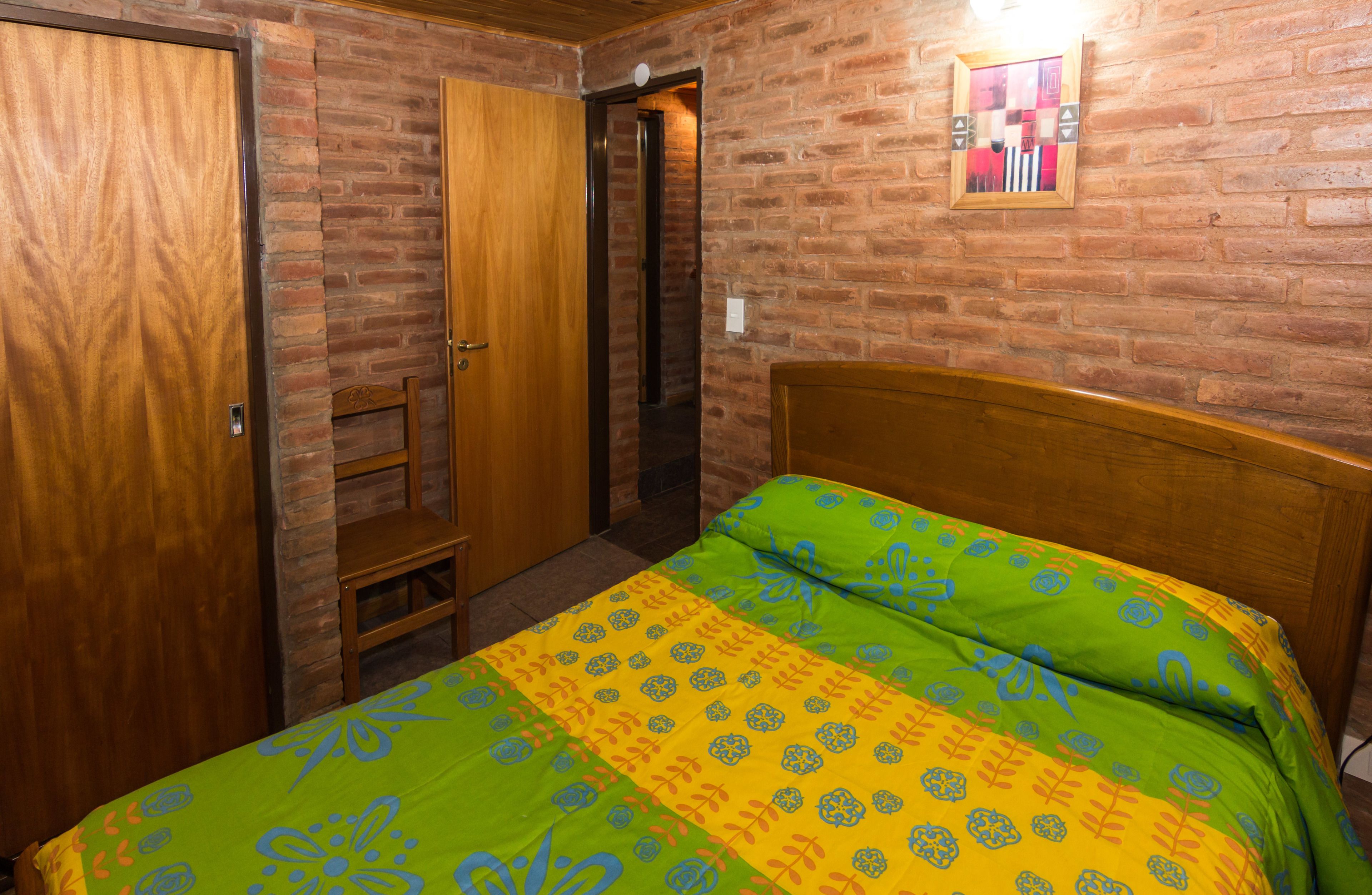 A bedroom with a brick wall, a wooden door, a bed with a patterned bedspread, and a small shelf.