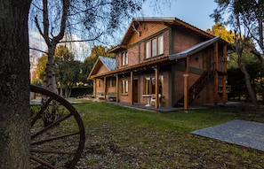 A rustic wooden house with a porch, a staircase, and a large tree in the foreground. - Los Maitenes Bariloche (San Carlos de Bariloche)