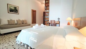 A hotel room with a bed, a sofa, a bookshelf, and a desk with a lamp.