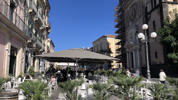 An outdoor café with tables and chairs, surrounded by potted plants and historic buildings.