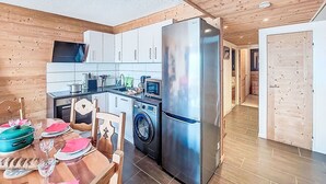 Une cuisine compacte avec des meubles blancs, un réfrigérateur en acier inoxydable, un lave-linge et un coin repas agrémenté d’une table en bois, mise pour deux personnes.