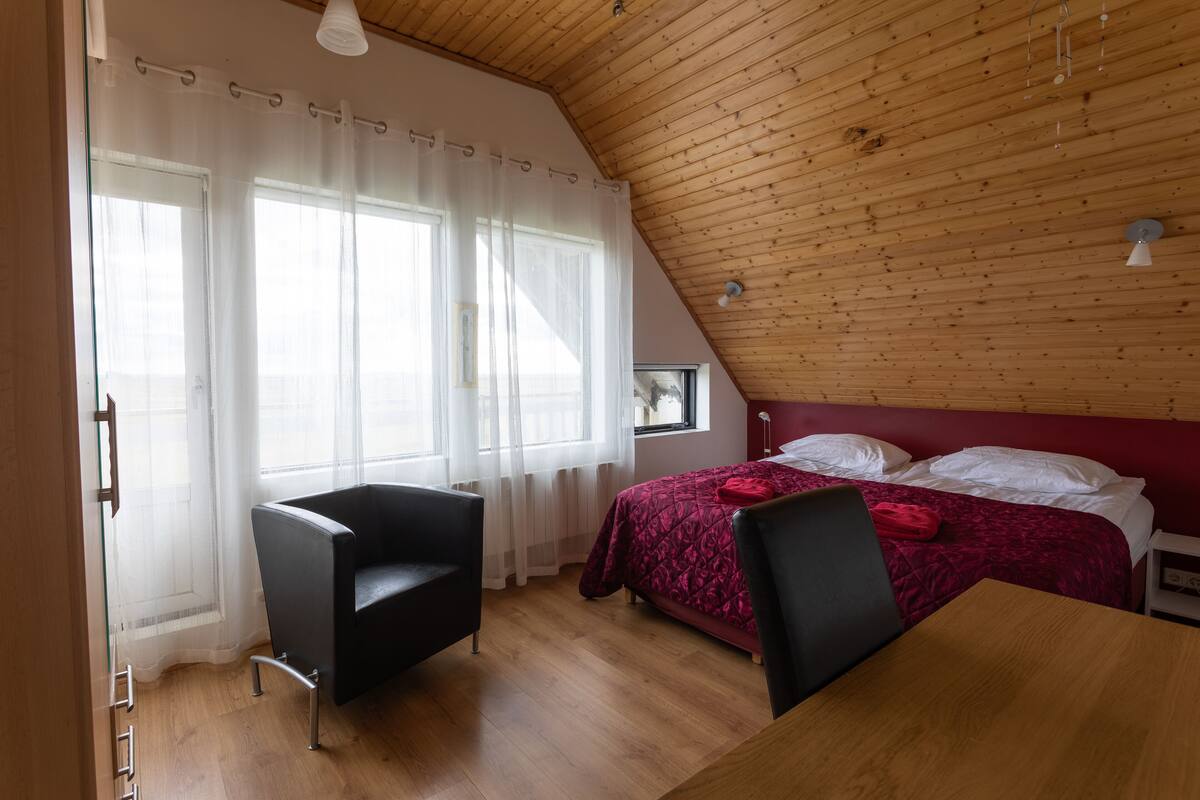 Un dormitorio con techo de madera, una cama con colcha roja, una silla negra y una mesa de madera.