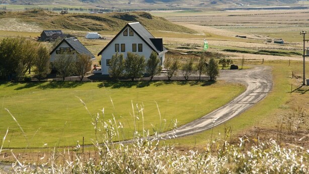 A rural landscape with two white houses, a dirt road, and rolling hills in the background. - Guesthouse Dalbaer (Hrunamannahreppur)