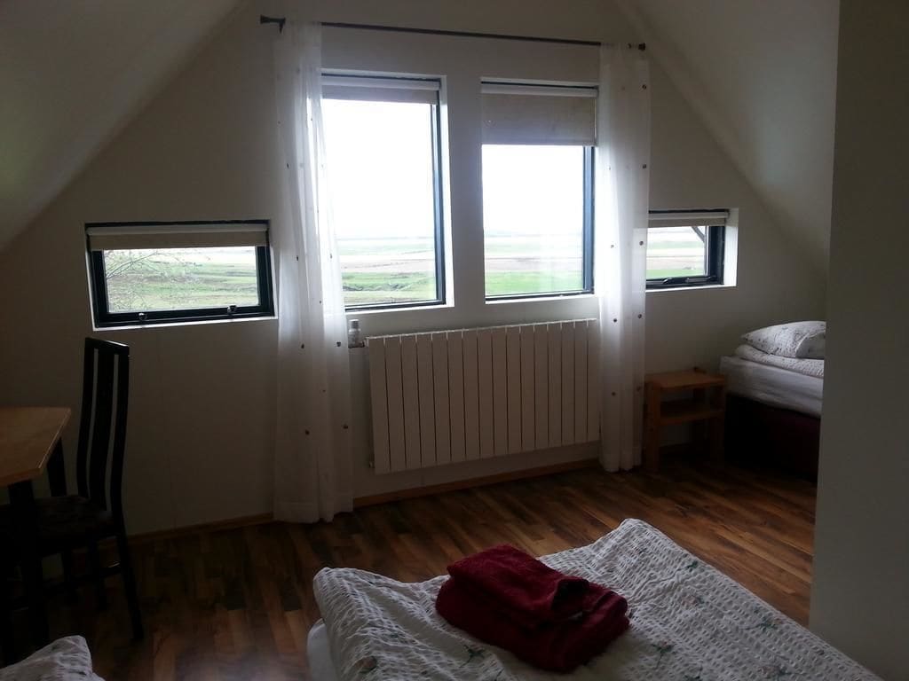 Une pièce avec un plafond en pente, deux fenêtres, un radiateur, un lit recouvert d’une couverture rouge et une petite table.