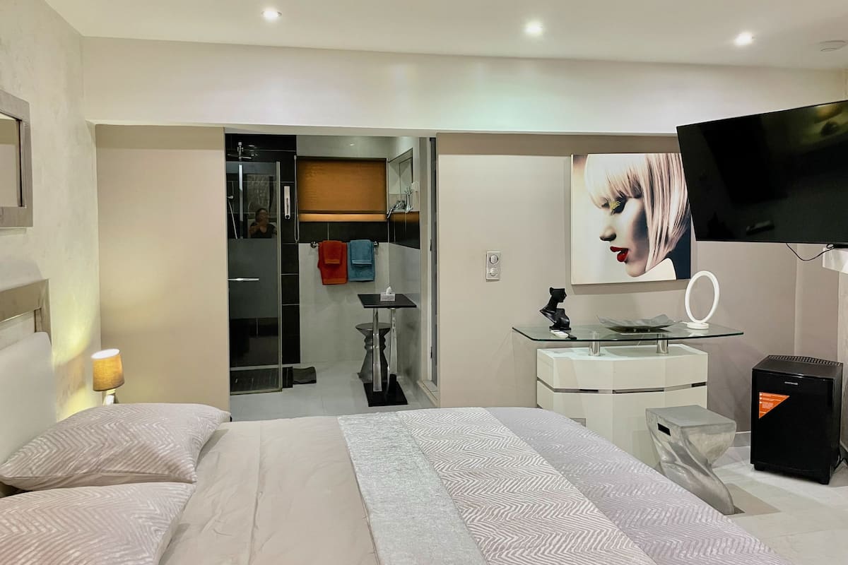 A modern hotel room with a large bed, a vanity with a mirror, a television, and a small refrigerator.