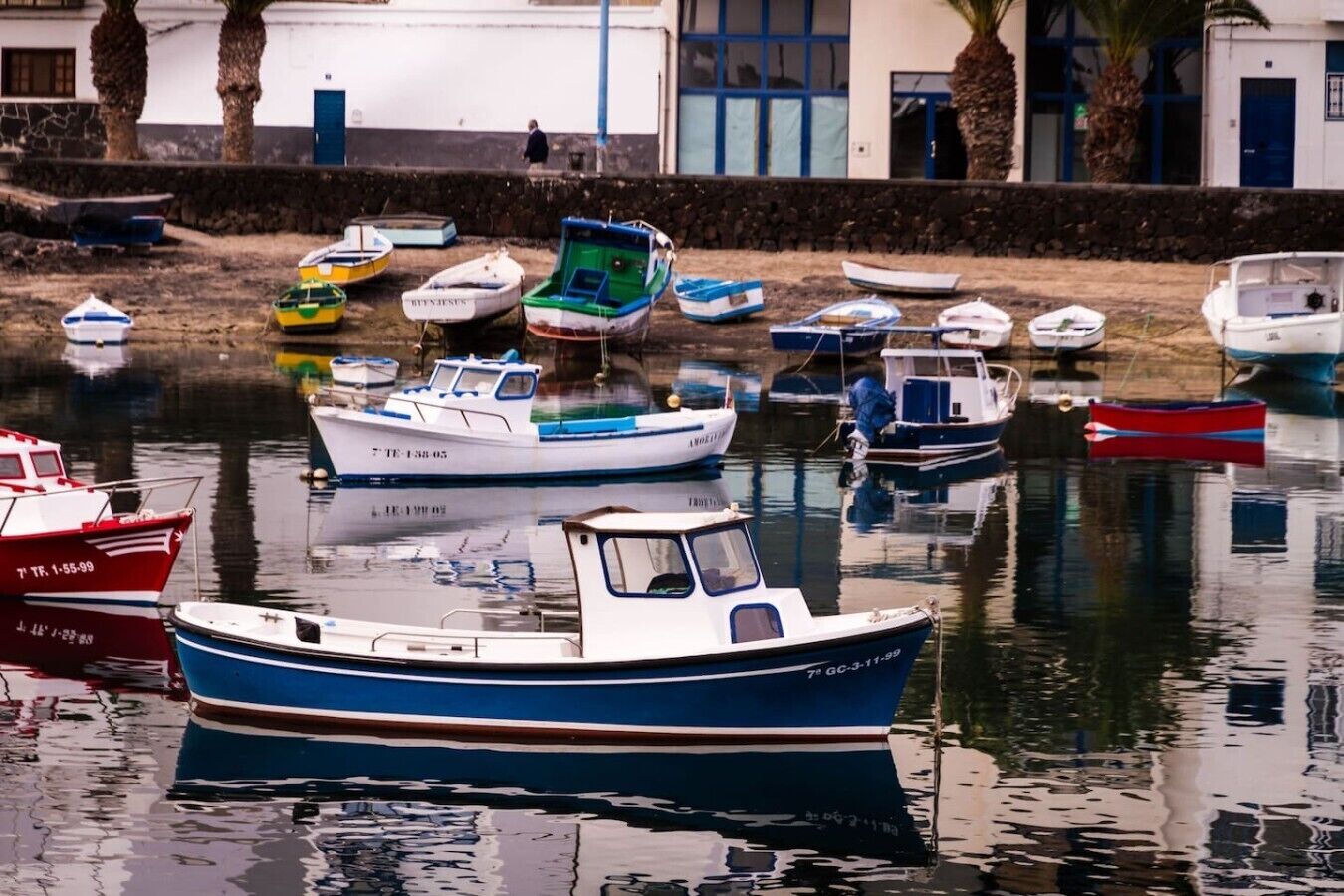 Un puerto deportivo con varias embarcaciones amarradas, una superficie de agua tranquila y un edificio con puertas azules al fondo.