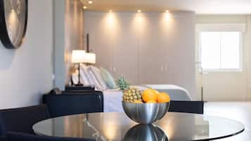 Una habitación de hotel moderna con comedor que presenta una mesa de vidrio redonda y sillas, un bol de metal con frutas, y una cama con un mesita de noche al fondo.