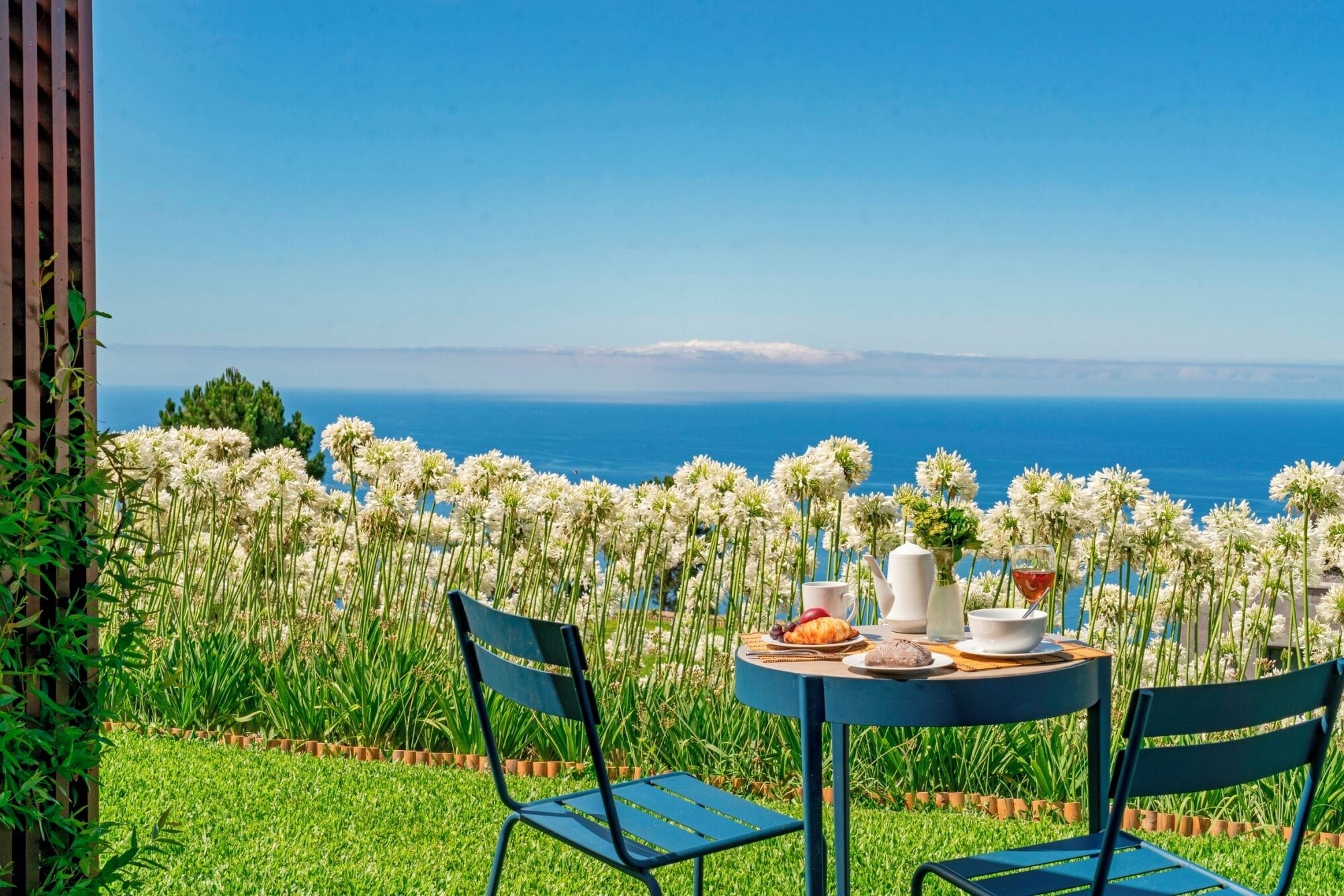Une table dressée avec un croissant, un bol, une tasse et un verre de vin rouge, donnant sur une vue sur la mer, avec des fleurs blanches au premier plan.
