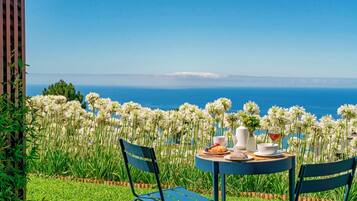 Une table dressée avec un croissant, un bol, une tasse et un verre de vin rouge, donnant sur une vue sur la mer, avec des fleurs blanches au premier plan.