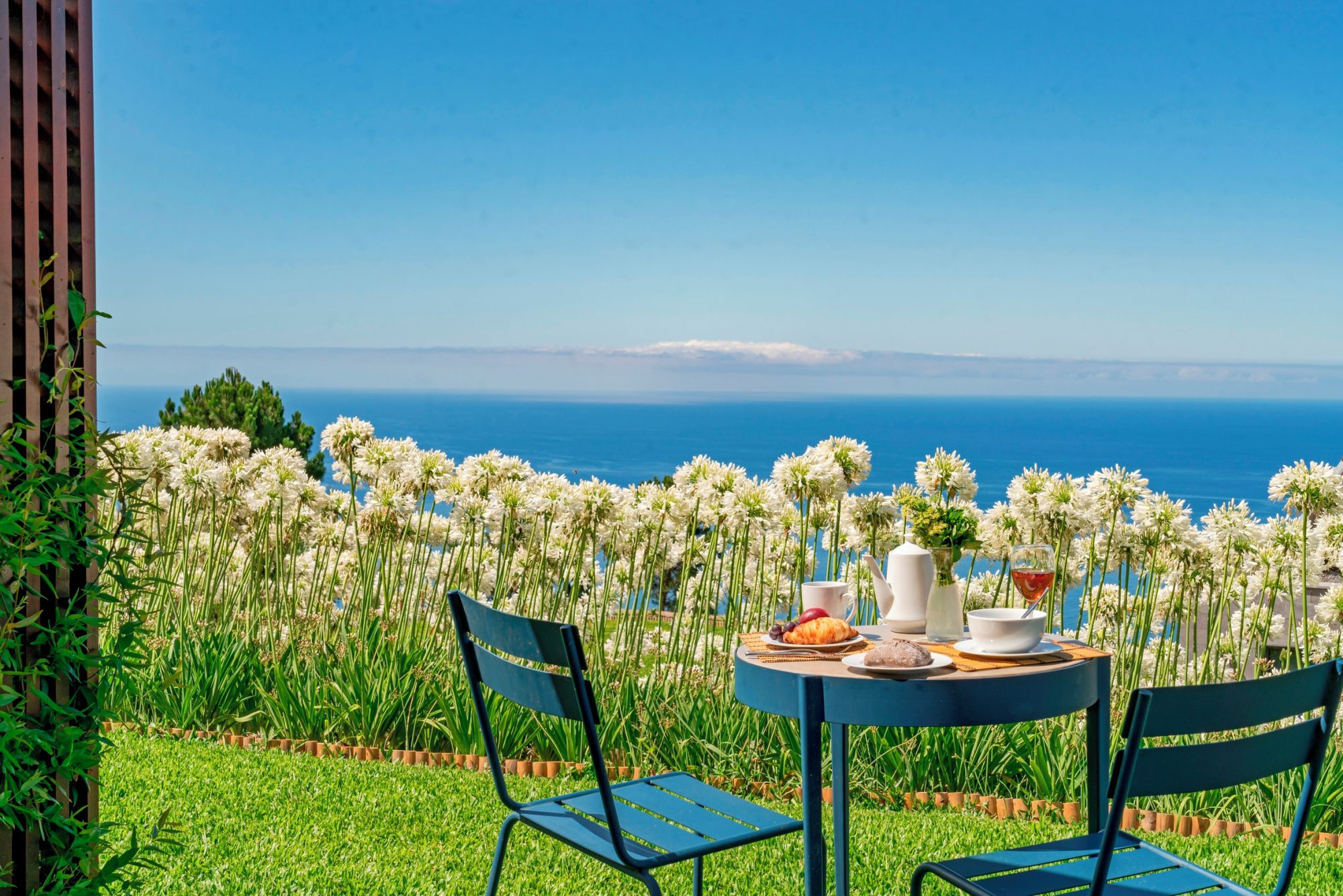 Et bord dækket med en croissant, en skål, en kop og et glas rødvin, med udsigt over havet og hvide blomster i forgrunden.