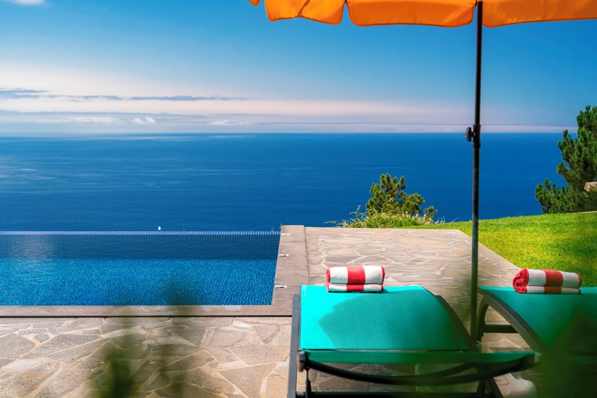 A pool with a view of the sea, a green lounge chair, and an orange umbrella.