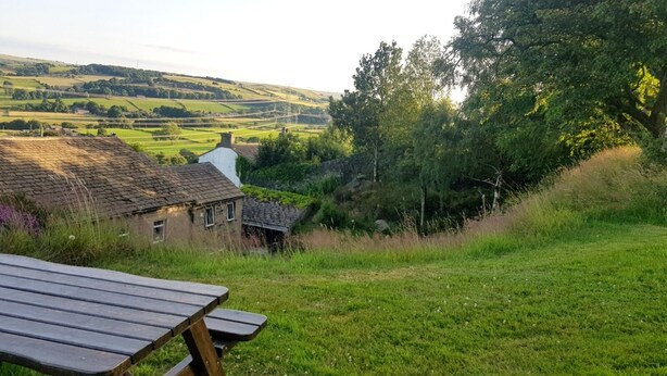 Une table de pique-nique se trouve dans un espace verdoyant, offrant une vue sur une maison et des collines ondulantes en arrière-plan.