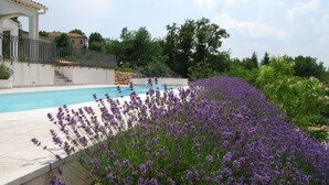Une piscine entourée de plantes de lavande, un bâtiment blanc agrémenté d’une pergola et un mur de pierre.