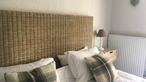 A bed with white bedding and plaid pillows, a wicker headboard, a bedside table with a lamp, and a radiator.