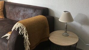 A brown sofa with a beige throw blanket, a small round wooden side table with a lamp, and a white textured wall.