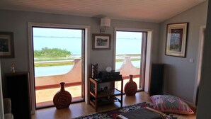 Une pièce dotée d’une grande porte-fenêtre coulissante donnant sur un balcon avec vue sur une étendue d’eau. On y trouve une console en bois sur laquelle sont disposés divers objets, un tapis sur le sol et un tableau encadré au mur.