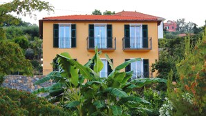 A two-story yellow building with a red roof, surrounded by lush greenery and a stone wall.