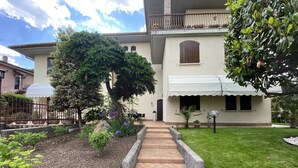 A courtyard with a paved pathway leading to a building with a balcony. There are potted plants and a small tree in the foreground.