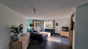 A modern living room with a sofa, coffee table, and TV stand. There is a cabinet with a TV on top, a ceiling fan, and a window with curtains. - Vacation apartment for 11 persons (incl. consumption costs and tourist tax!) (Zeewolde)