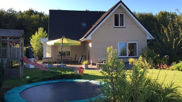 Ein Haus mit Backsteinfassade, einem Ziegeldach und einem Vorgarten mit Trampolin und einer hölzernen Spielanlage.