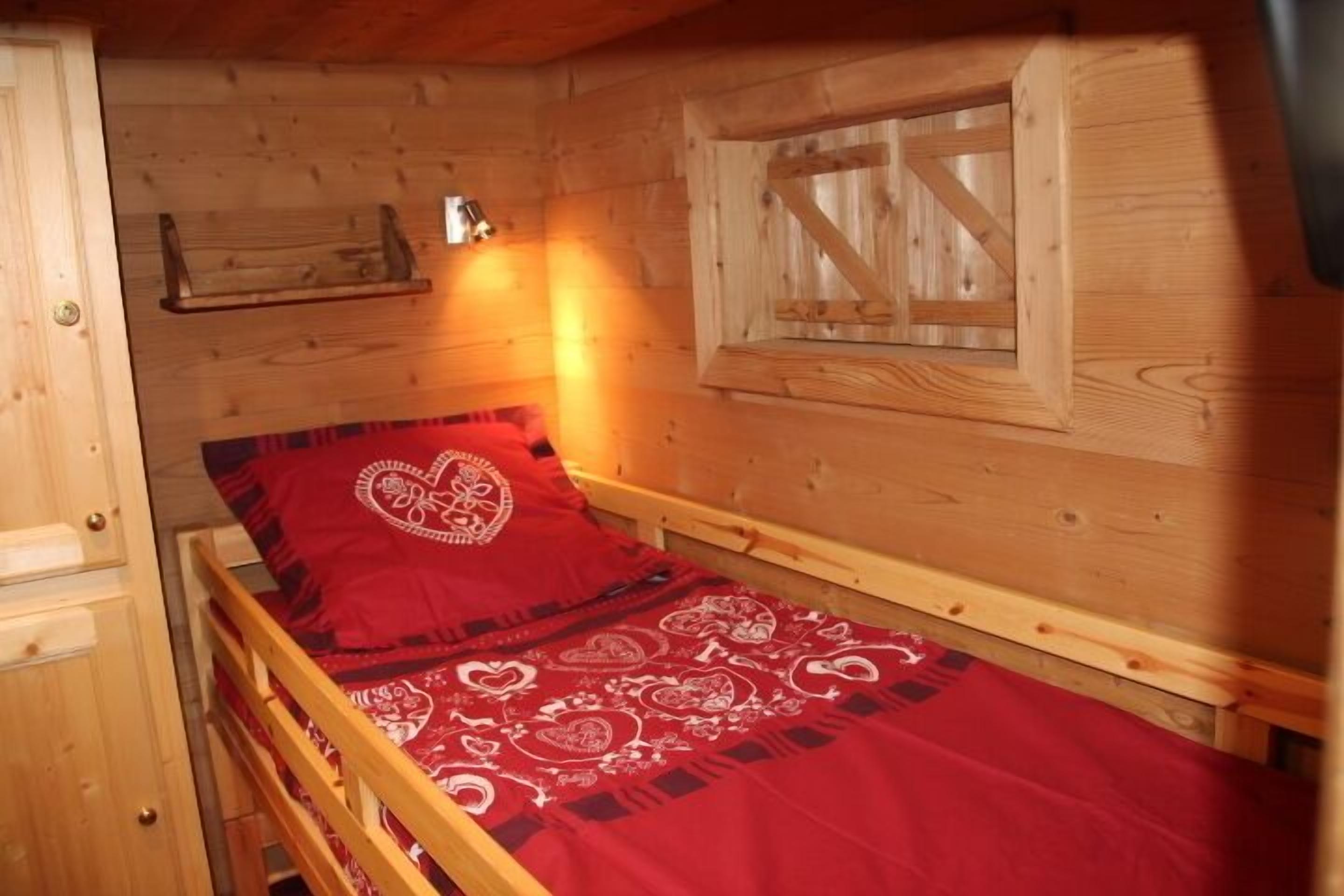 A wooden cabin room with a single bed, a window with wooden shutters, and a small shelf above the bed.