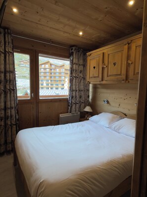 A bedroom with a wooden headboard, a bed with white linens, a nightstand with a lamp, and a window with curtains. - Studio - VAL THORENS (Les Belleville)