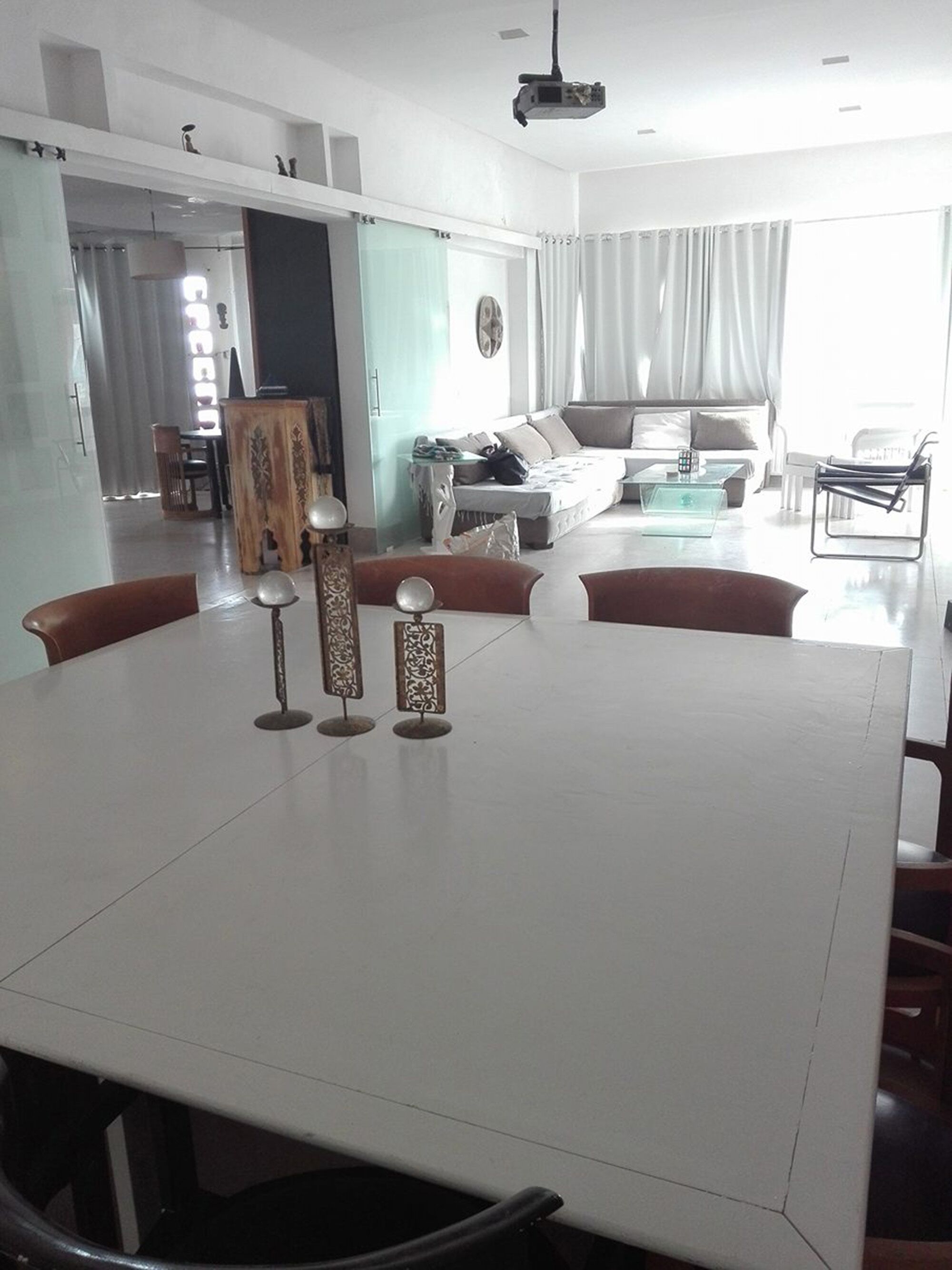 Een moderne eetruimte met een witte tafel en stoelen, een beamer, en een uitzicht op een woonkamer met een bank en gordijnen.