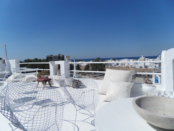 Terraza en la azotea con mobiliario blanco, que incluye una mesa redonda y una silla de metal, con vistas a un paisaje urbano costero.