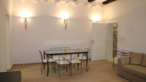 Un área de comedor con una mesa de vidrio y sillas, un sofá, y luces fijadas a la pared.