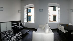 Una sala de estar moderna con un sofá, una mesa de comedor y un televisor. Hay ventanas con contraventanas y un aparador de vidrio.